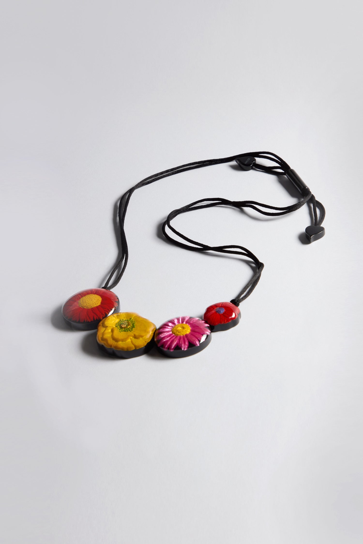 The Garland Necklace is a vibrant statement piece featuring a black cord with four round, hand-painted floral pendants on a white background.