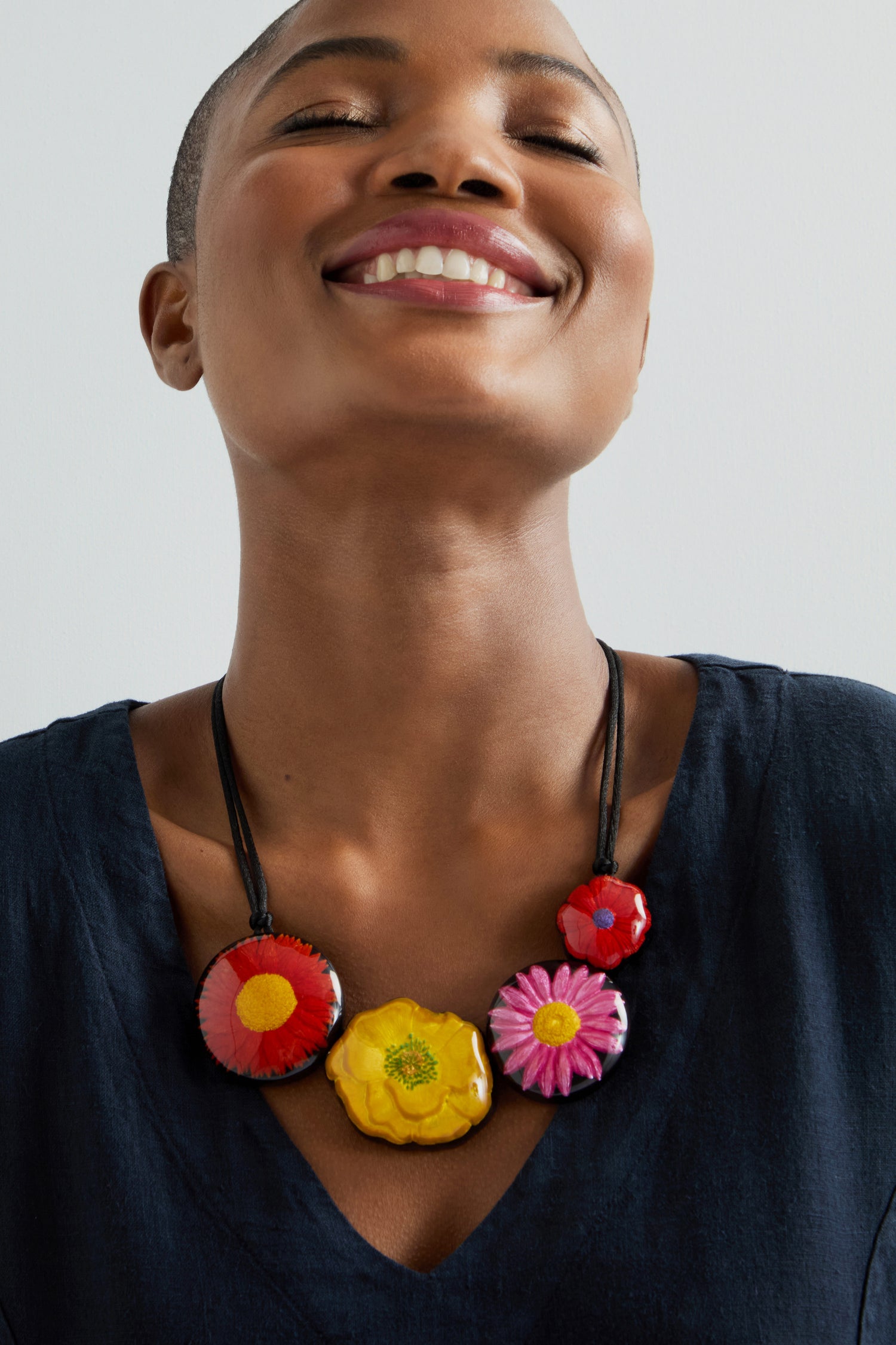 A person smiles wearing the Garland Necklace, a vibrant statement piece of hand-painted jewelry that exudes floral charm, paired beautifully with a dark V-neck top.