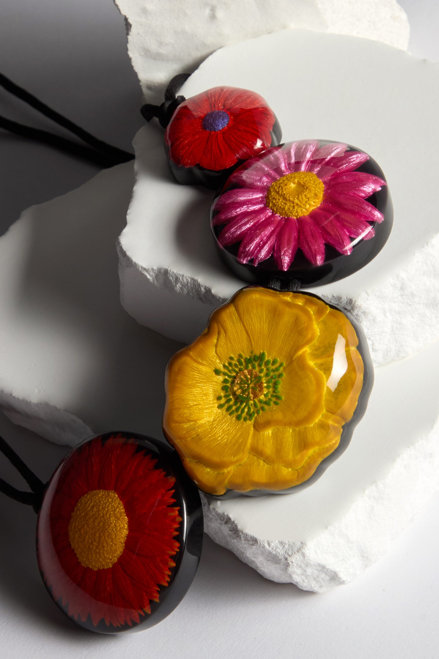 Explore the Garland Necklace featuring circular pendants with realistic, hand-painted red, pink, and yellow flower designs. Displayed on textured white surfaces, these resin necklaces are perfect for adding a vibrant pop of color to any outfit.