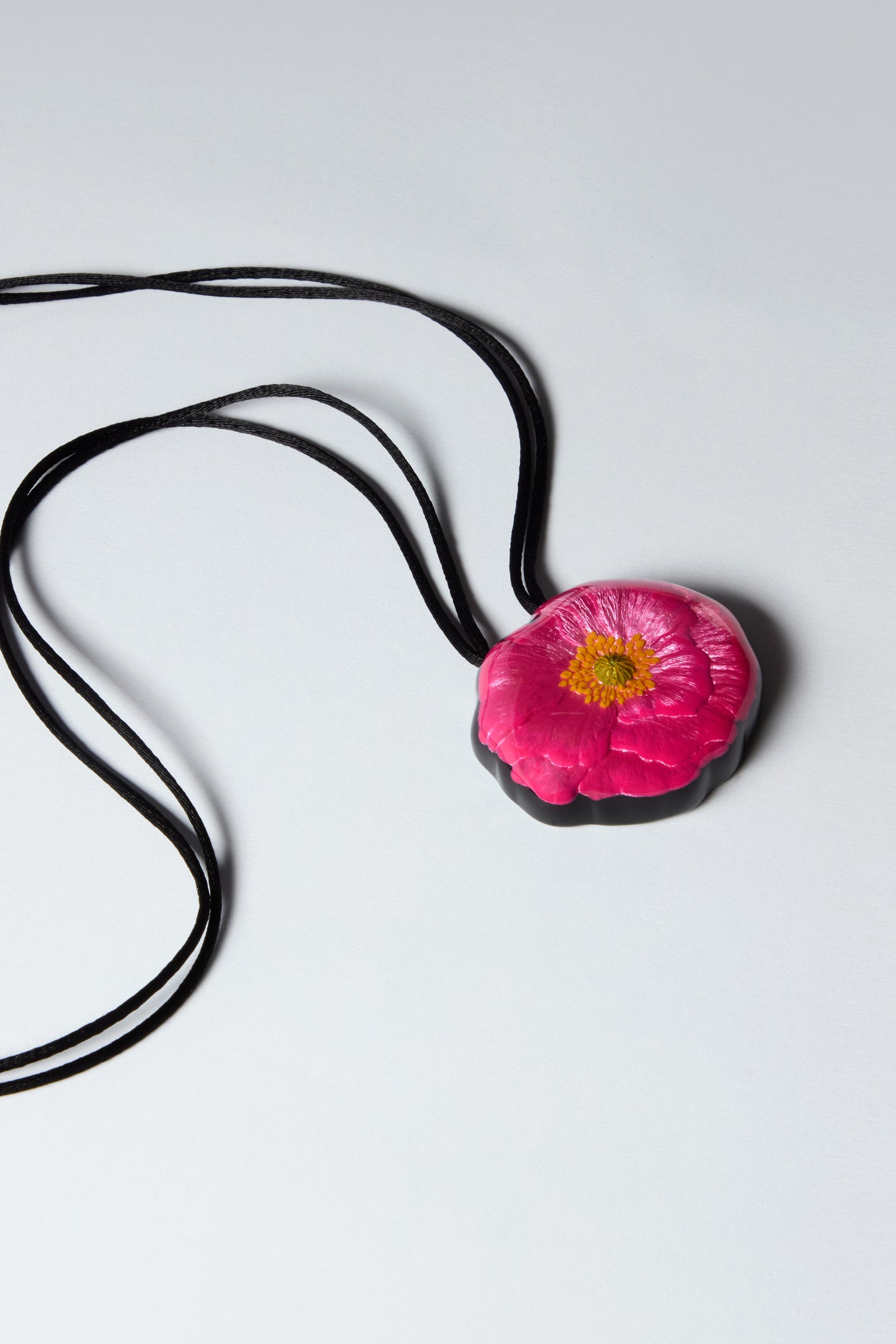 The Garland Pendant, a handcrafted pink flower design, delicately rests on an adjustable black cord necklace against a serene light gray background.