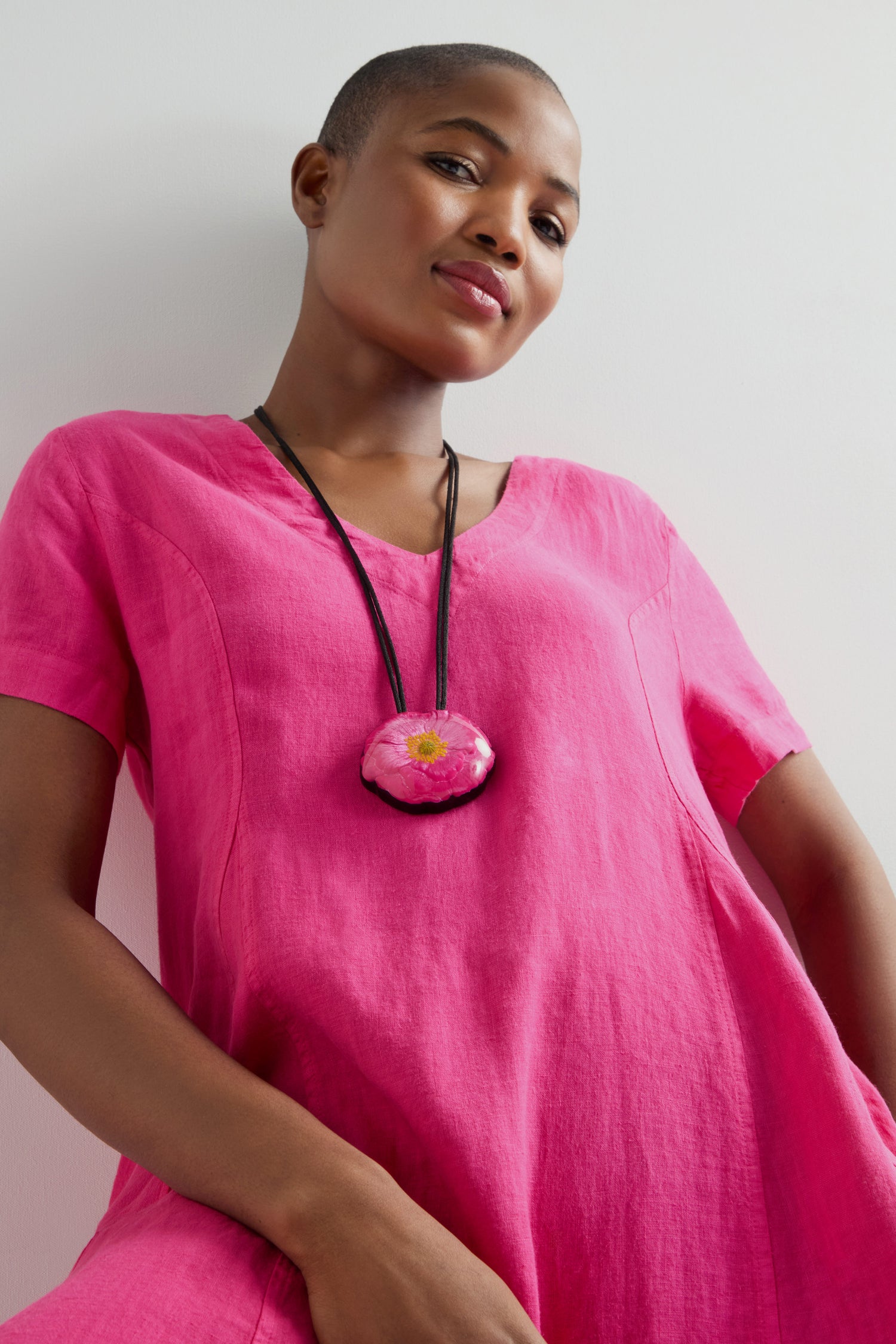 Wearing a vivid pink dress, someone pairs it with the Garland Pendant—an elegant black necklace featuring a handcrafted round floral design. The necklace is set against a light backdrop and includes an adjustable cord for maximum comfort and fit.