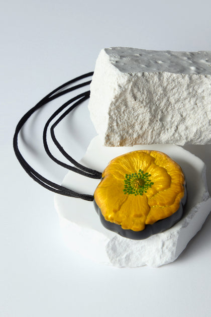 The Garland Pendant, a handcrafted yellow flower with adjustable black cords, gracefully rests on textured white stones.