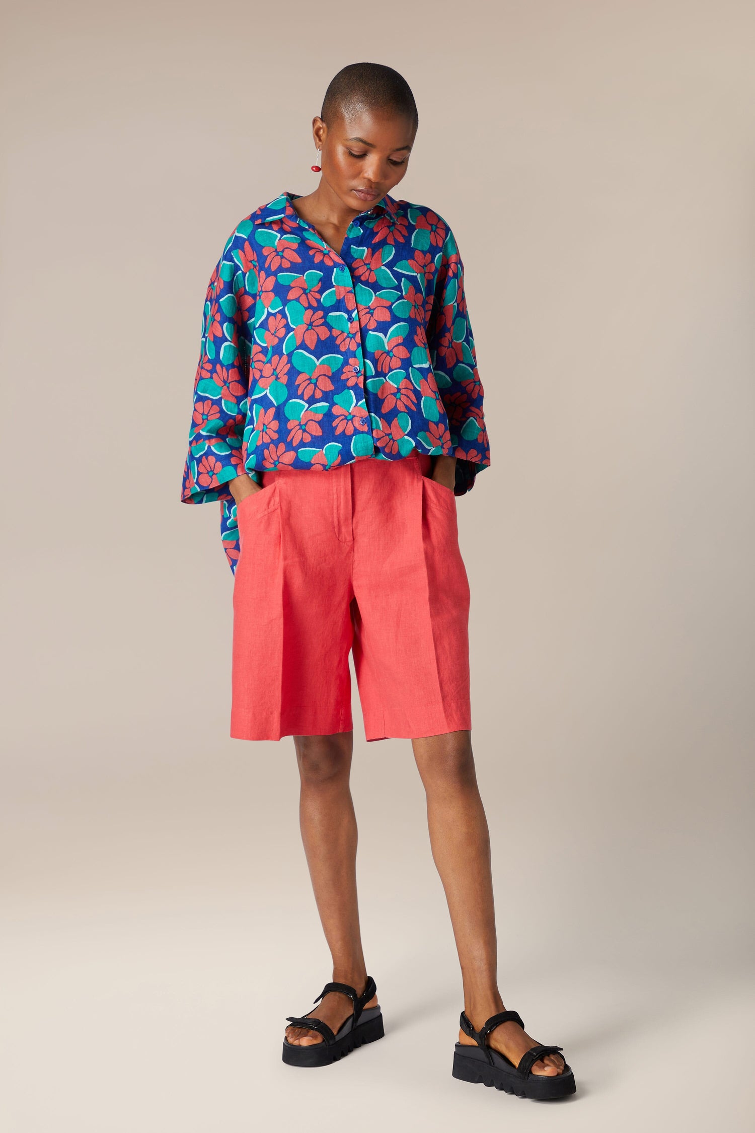 A person stands in a studio, wearing a vibrant blue floral shirt, [Bradley Linen Shorts], and black platform sandals, with a neutral background.