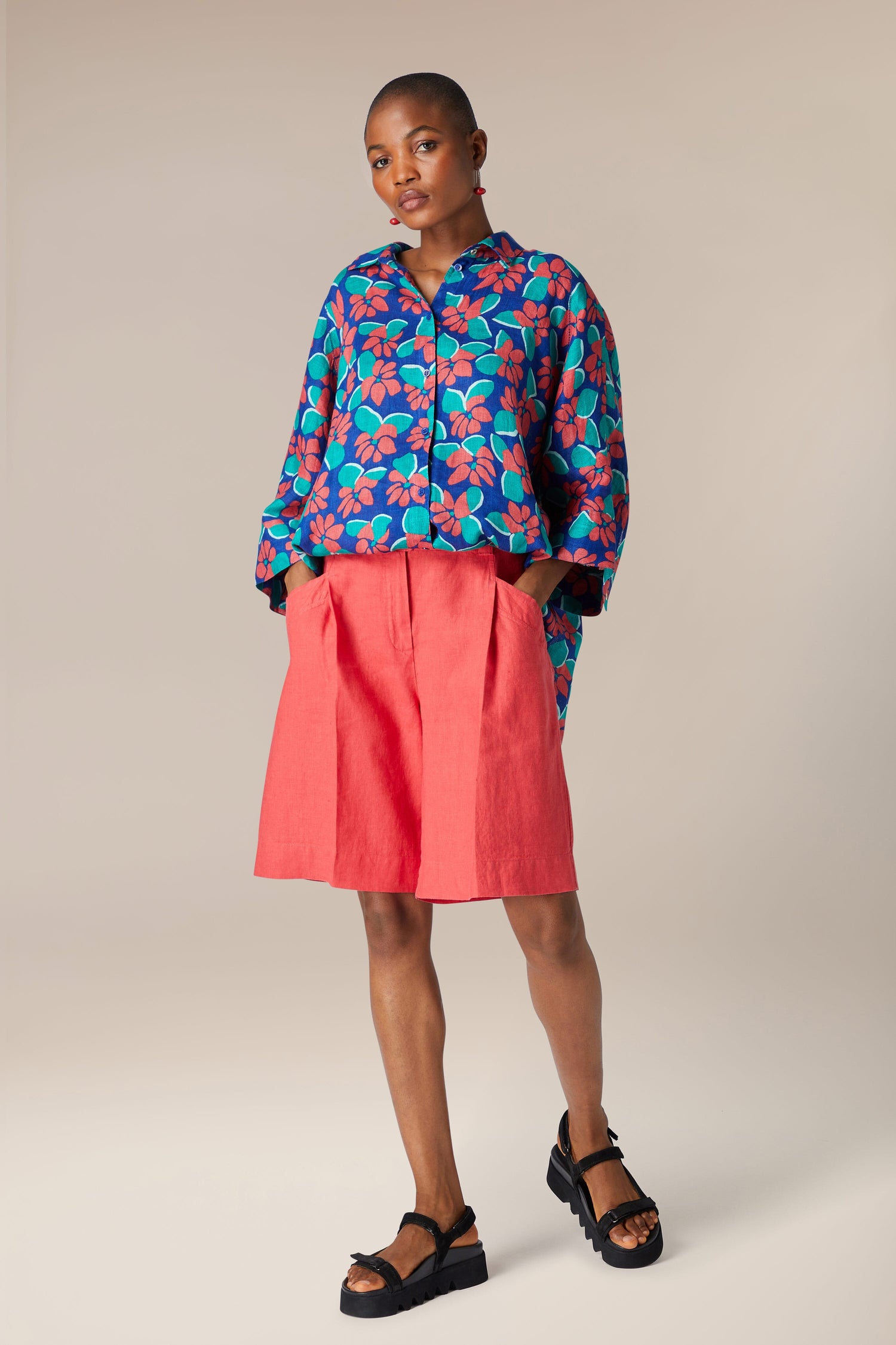A model stands wearing a floral blue shirt, Bradley Linen Shorts, and black sandals against a neutral background.
