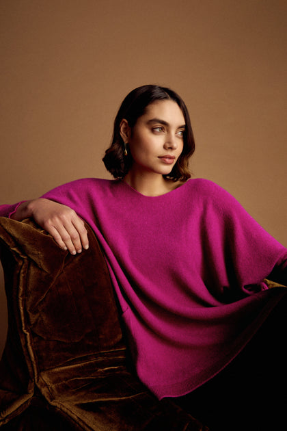 A woman with dark hair and gold earrings sits on a brown velvet chair, wearing the Two Pocket Oversize Knit in bright magenta, against a plain brown background.