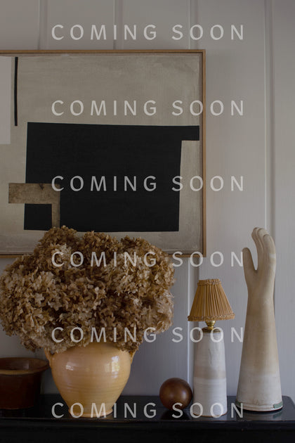 A beige vase with dried flowers, a lamp, a decorative hand, and a wooden ball are arranged on a surface in front of a black and beige abstract painting. The scene exudes the same elegance as the Cashmere Blend Crop Jumper. "COMING SOON" text is repeated across the image.