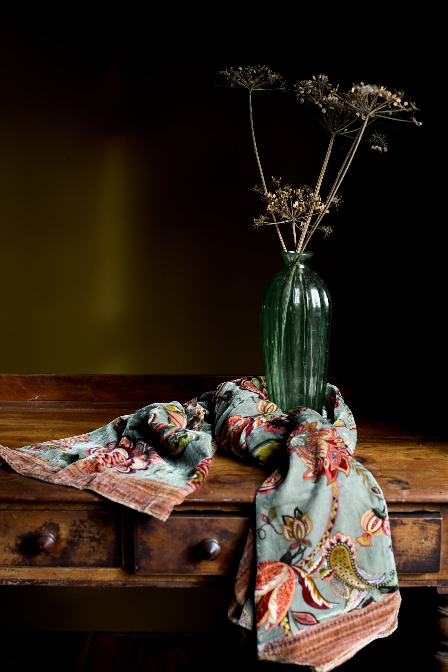A green glass vase with dried flowers sits on a wooden table beside the In Bloom Velvet Scarf, folded to showcase its earthy patterns—a subtle statement accessory for any room.