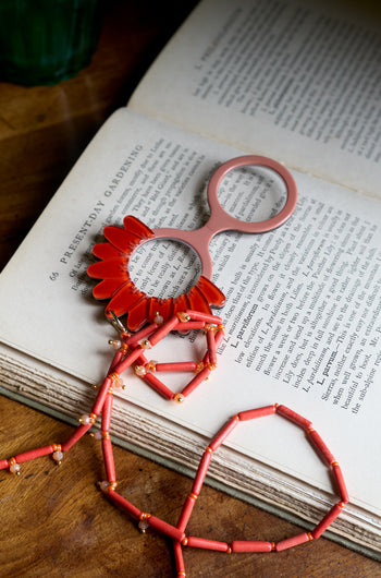A pair of Daisy Pendant Glasses with a pink floral edge rests on the open pages of a book atop a wooden table.