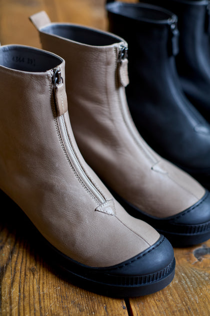 A close-up of the Biscotti Zip Front Leather Boots highlights handcrafted beige and black Italian leather with front zippers and a chunky serrated sole, displayed on a wooden floor.