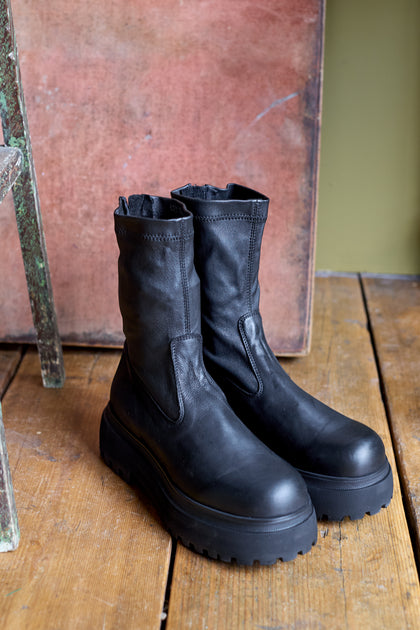 A pair of Short Stretch Leather Black Boots featuring handcrafted Italian leather and chunky platform soles sits on a wooden floor beside a rustic chair and textured background.