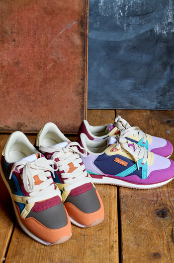 Two pairs of Colour Pop Andree Trainers are placed side by side on a wooden floor, set against a textured backdrop with brown and blue panels.