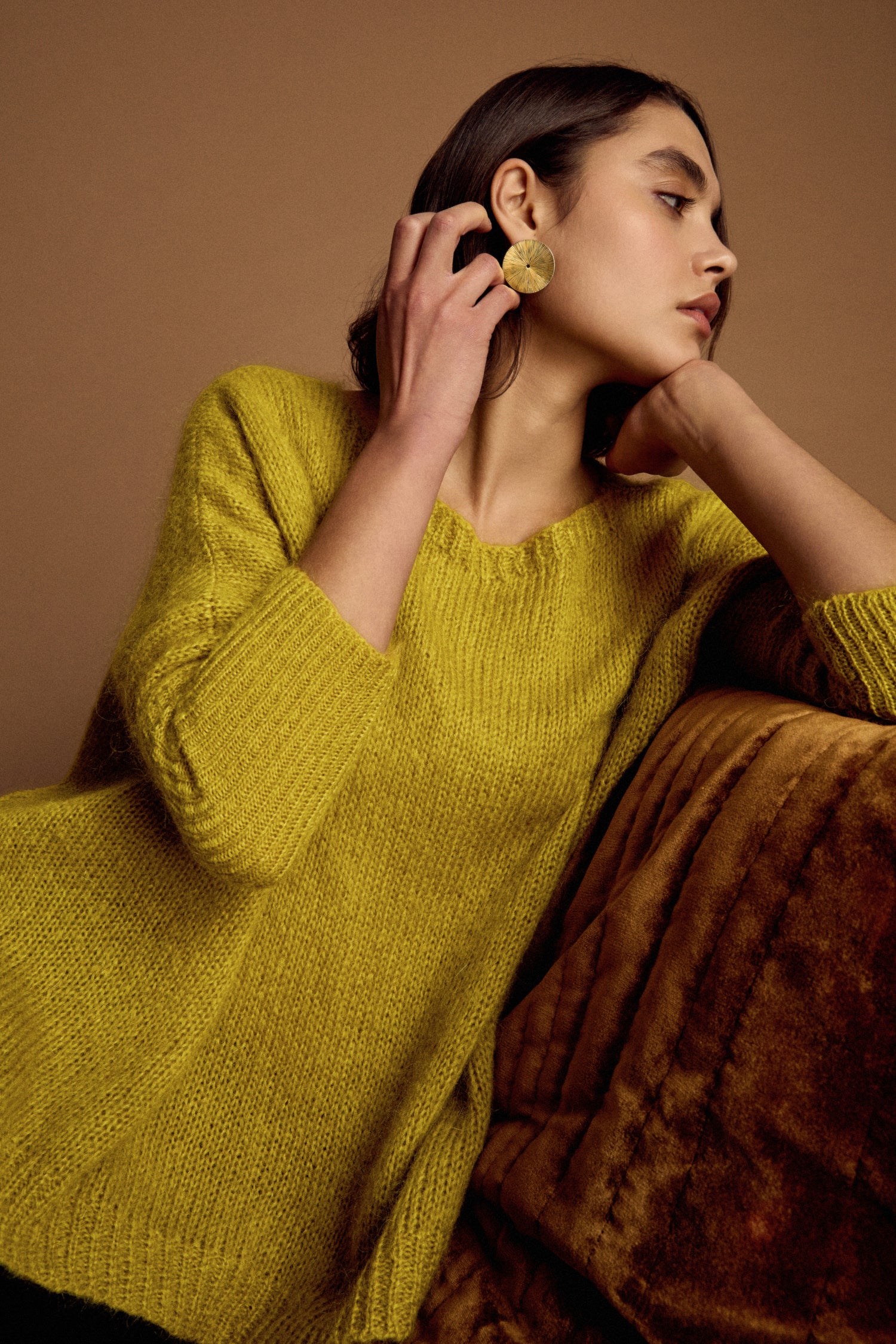 A woman in a Wide Neck Mohair Knit sweater and large round earrings sits on a brown sofa, resting her chin on her hand and gazing to the side.