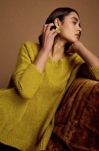 A woman in a Wide Neck Mohair Knit sweater and large round earrings sits on a brown sofa, resting her chin on her hand and gazing to the side.