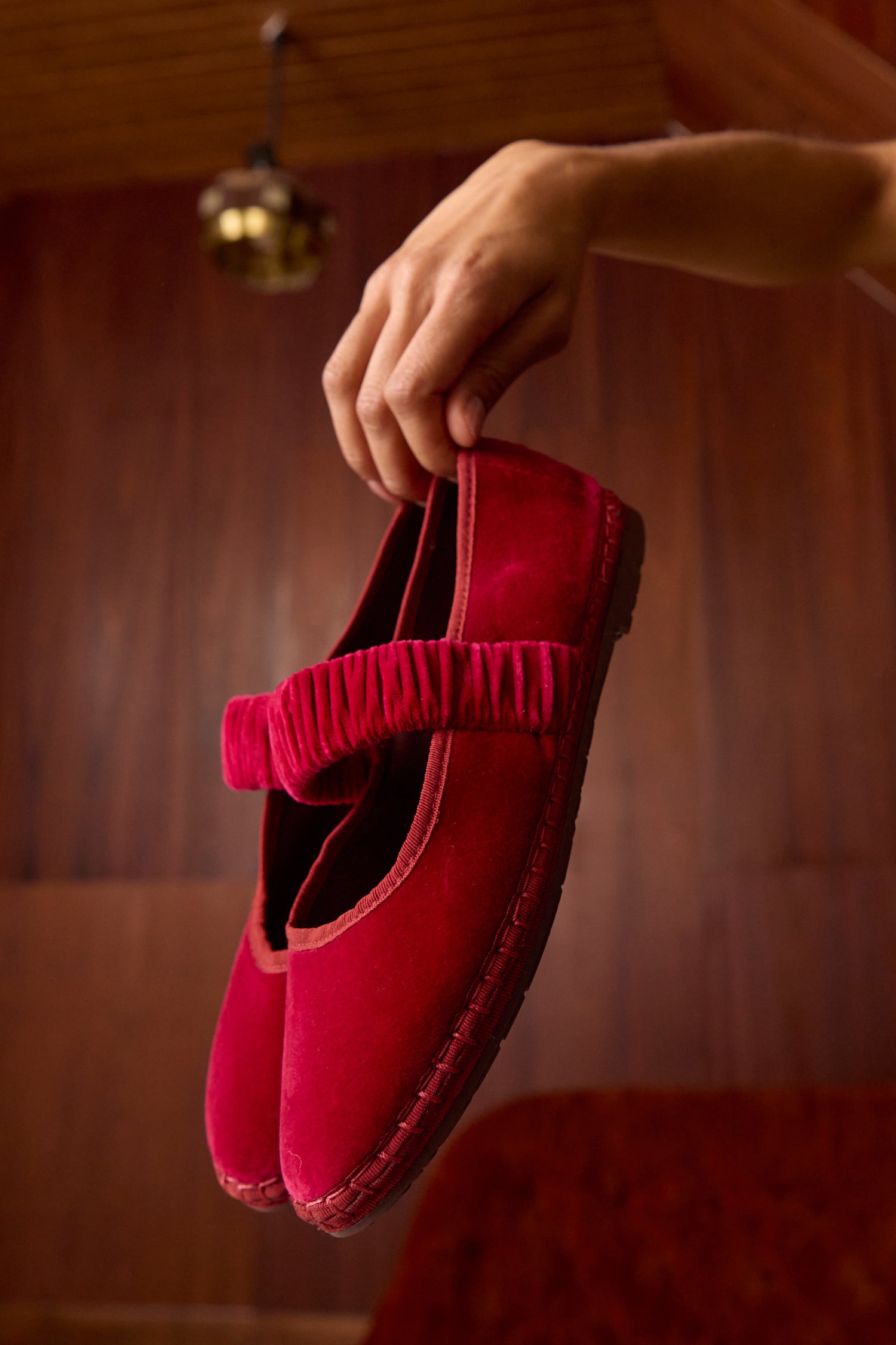 A hand holds the Mafalda Fuchsia Velvet Shoes, Mary Jane-style with ruched straps and handmade in Spain, set against a wooden background.