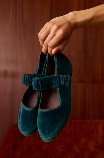 A hand holds up Mafalda Teal Velvet Shoes—handmade Mary Jane flats featuring a ruched strap and recycled rubber sole—against a wooden background.