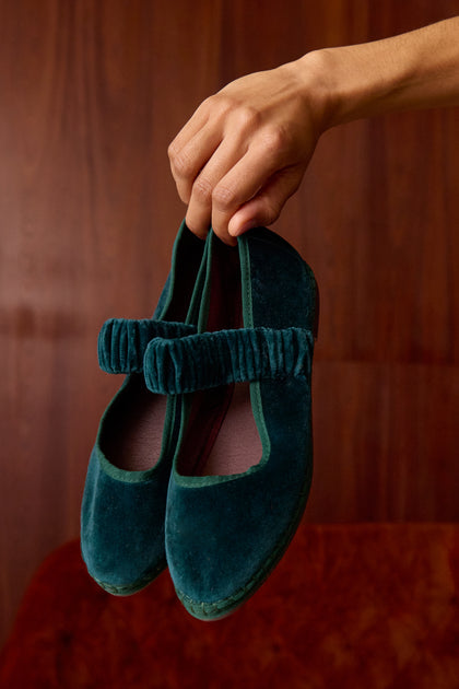 A hand holds up Mafalda Teal Velvet Shoes—handmade Mary Jane flats featuring a ruched strap and recycled rubber sole—against a wooden background.