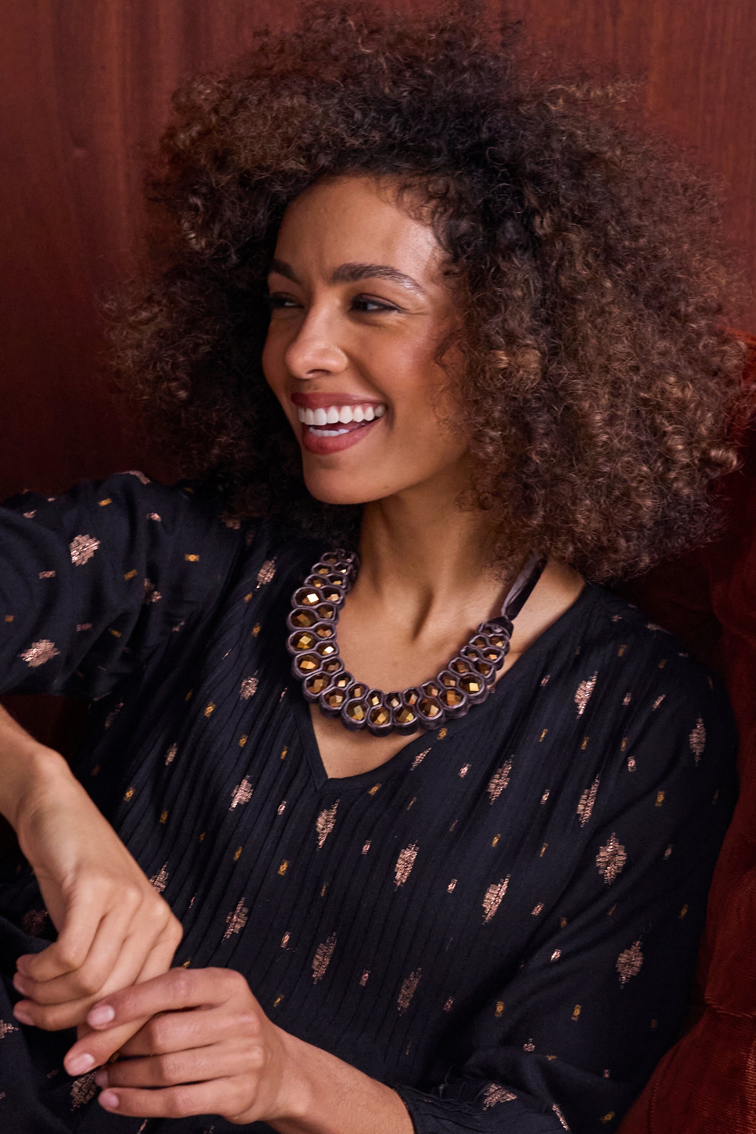 A woman with curly hair, in a black patterned top and Velvet Ribbon Statement Necklace, smiles while seated against a wooden background.