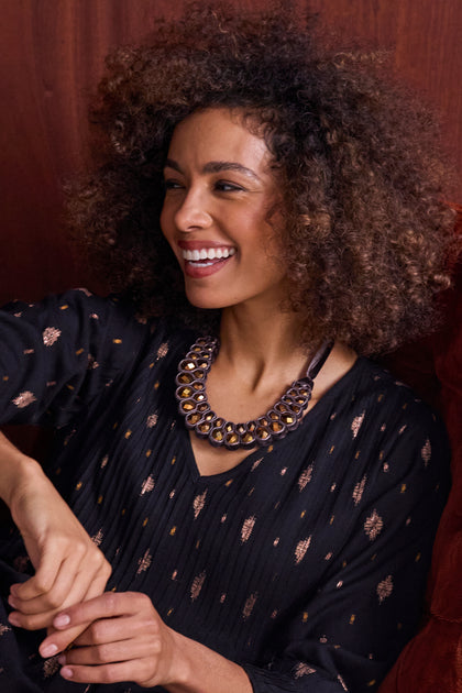 A woman with curly hair, in a black patterned top and Velvet Ribbon Statement Necklace, smiles while seated against a wooden background.