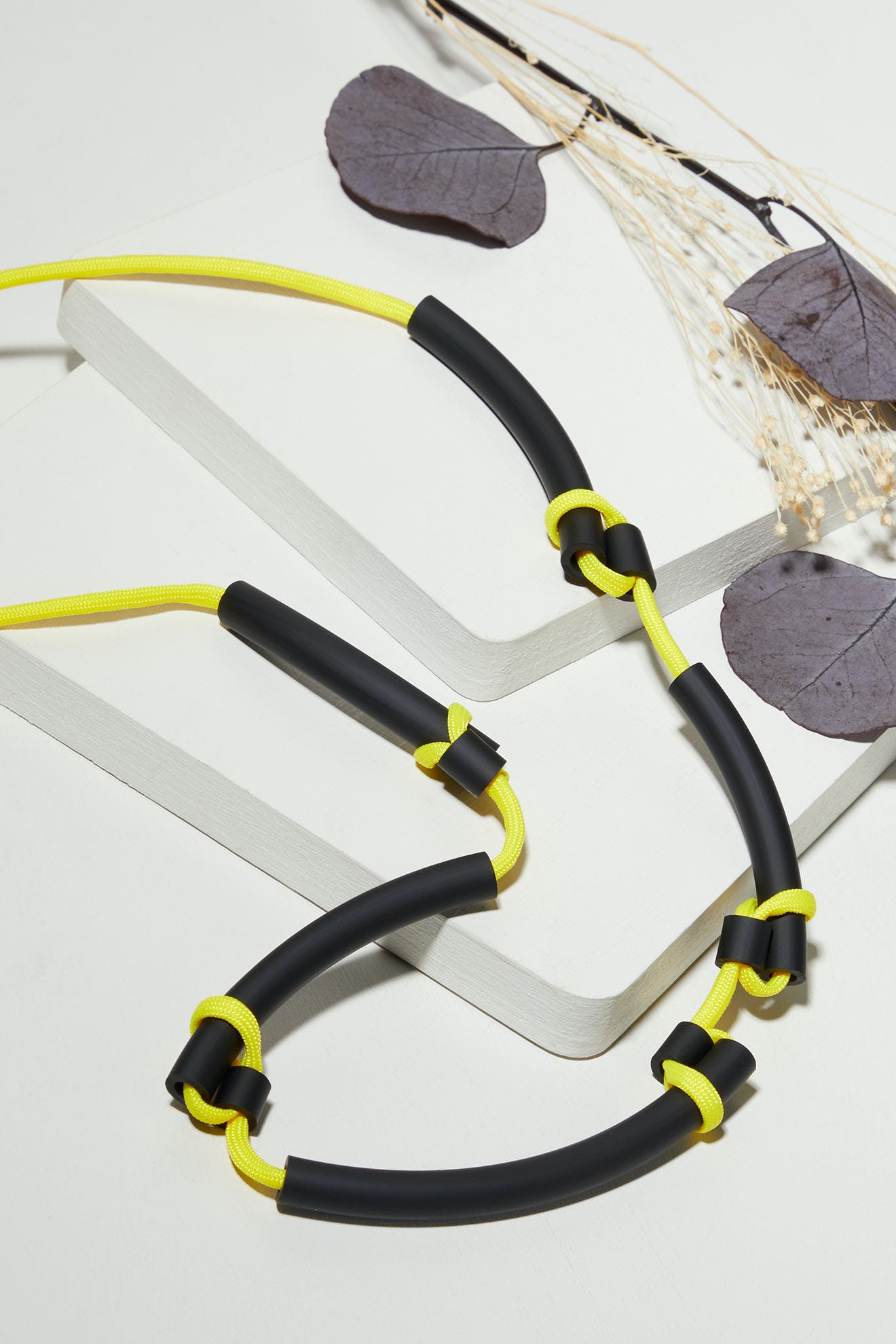 A Twists Necklace lies neatly coiled on a white surface, its vibrant yellow and black design accompanied by dried leaves and stems, evoking the distinctive craftsmanship of Samuel Coraux's bright jewelry collection.