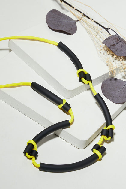 A Twists Necklace lies neatly coiled on a white surface, its vibrant yellow and black design accompanied by dried leaves and stems, evoking the distinctive craftsmanship of Samuel Coraux's bright jewelry collection.