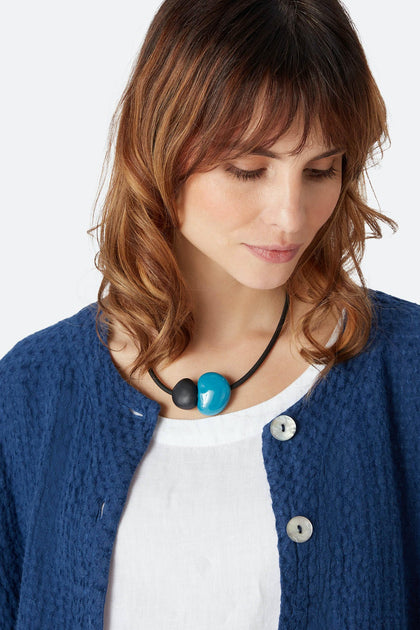 A woman with shoulder-length brown hair wears a textured blue jacket, a white top, and the Pebbles Necklace featuring round black and blue geometric beads, looking down with a neutral expression.