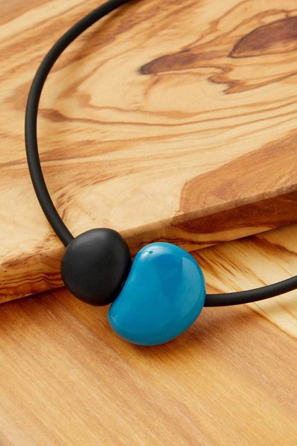 The Pebbles Necklace from Andreani Creations features a black cord with two beads—one black and one large, blue, and irregularly shaped—displayed on wood. Its bold geometric design is perfect for fans of handmade jewellery.