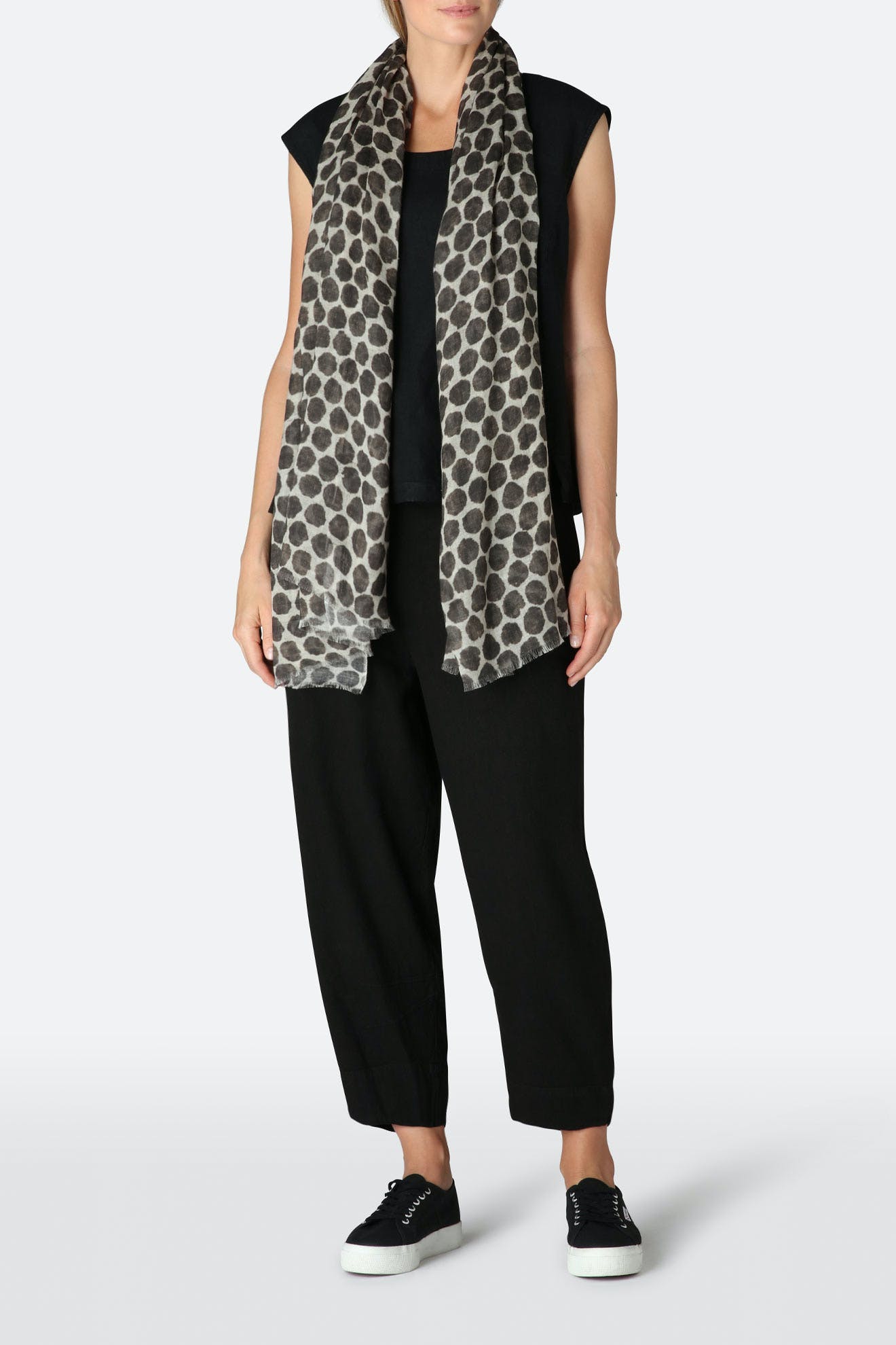 Person wearing a black sleeveless top, black pants, black sneakers, and the Circle Spot Scarf—a long lightweight accessory with a grey and black dot pattern—standing against a plain light background.