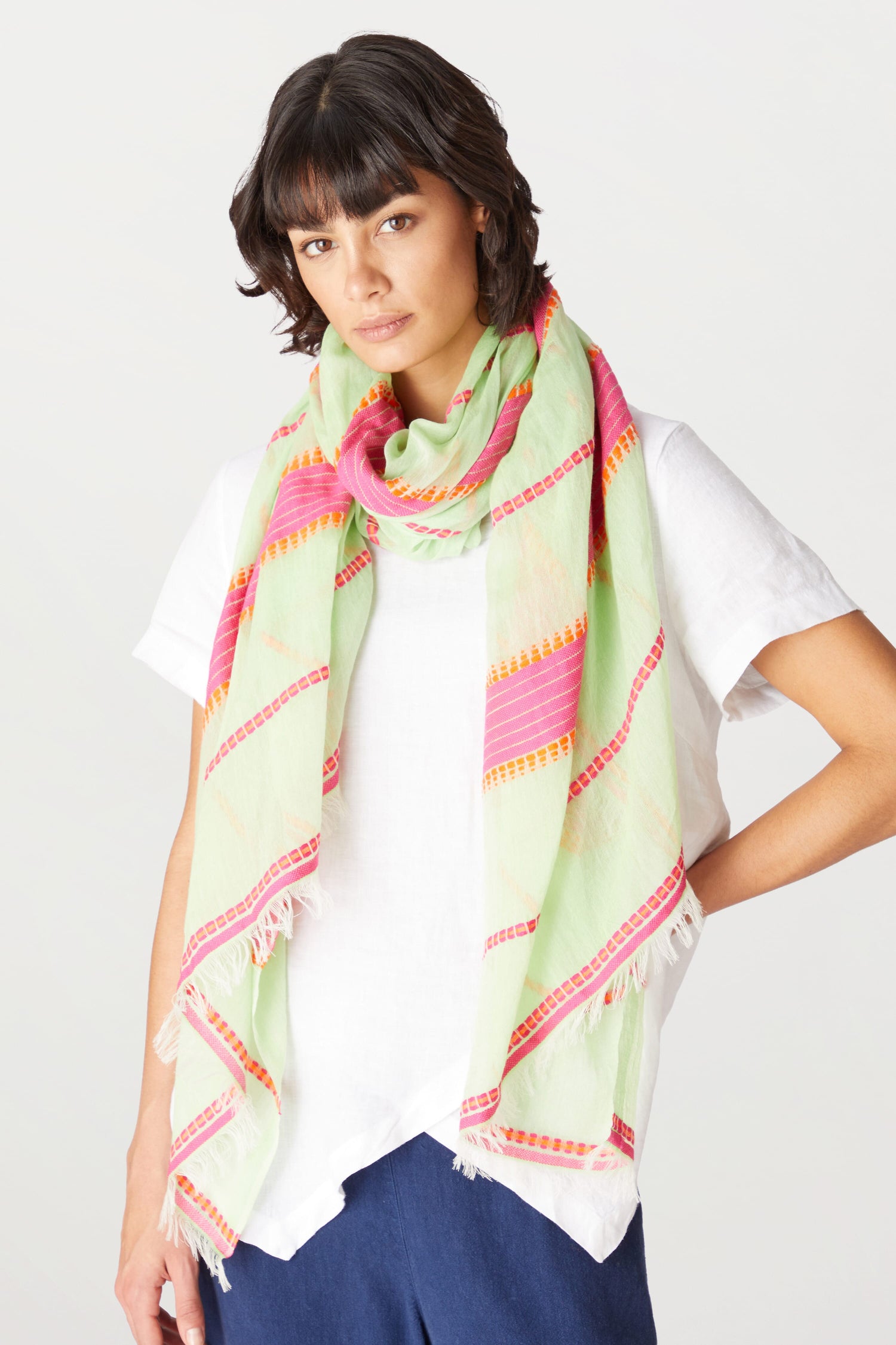 A woman with dark hair wearing a white top and a colorful Embroidered Stripe Scarf, standing against a light background.