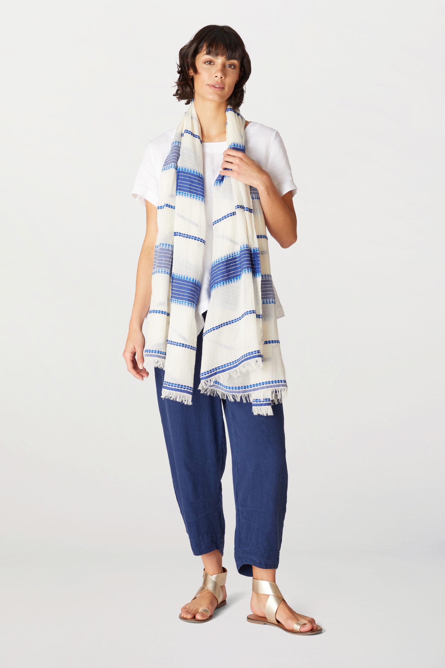 A woman in a white t-shirt, blue cropped pants, and white sandals, standing with an Embroidered Stripe Scarf draped around her neck.
