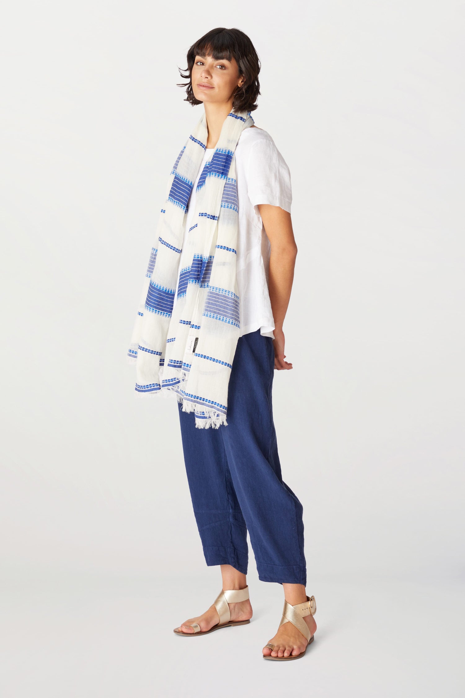 Woman in a white blouse, blue cropped trousers, and beige sandals posing with an Embroidered Stripe Scarf draped over one shoulder, against a light background.