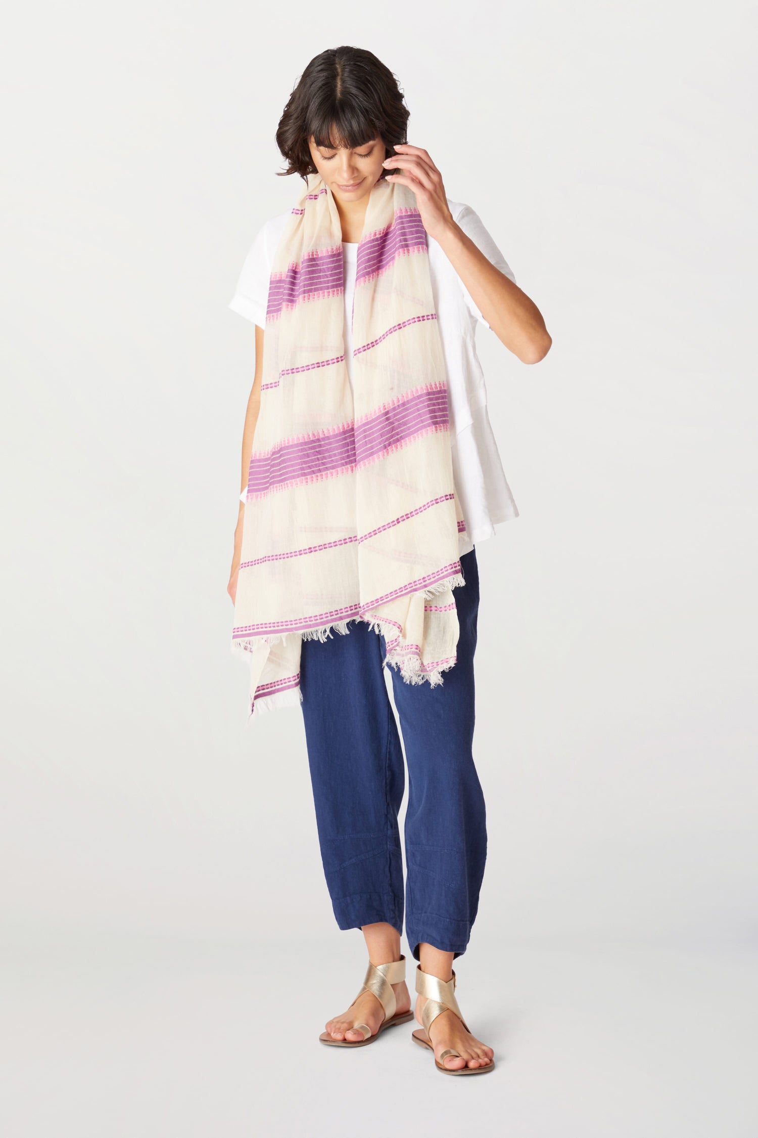 A woman standing in a studio, wearing a white top, distressed jeans, sandals, and a long Embroidered Stripe Scarf.