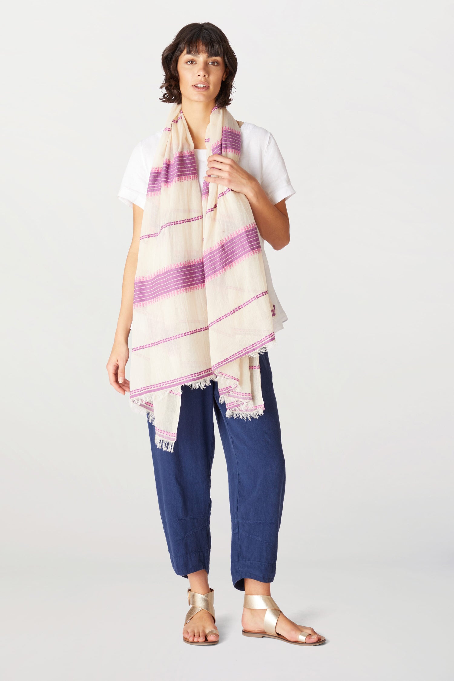 A woman stands wearing navy blue trousers, a white shirt, and an Embroidered Stripe Scarf, paired with gold sandals. She looks directly at the camera.