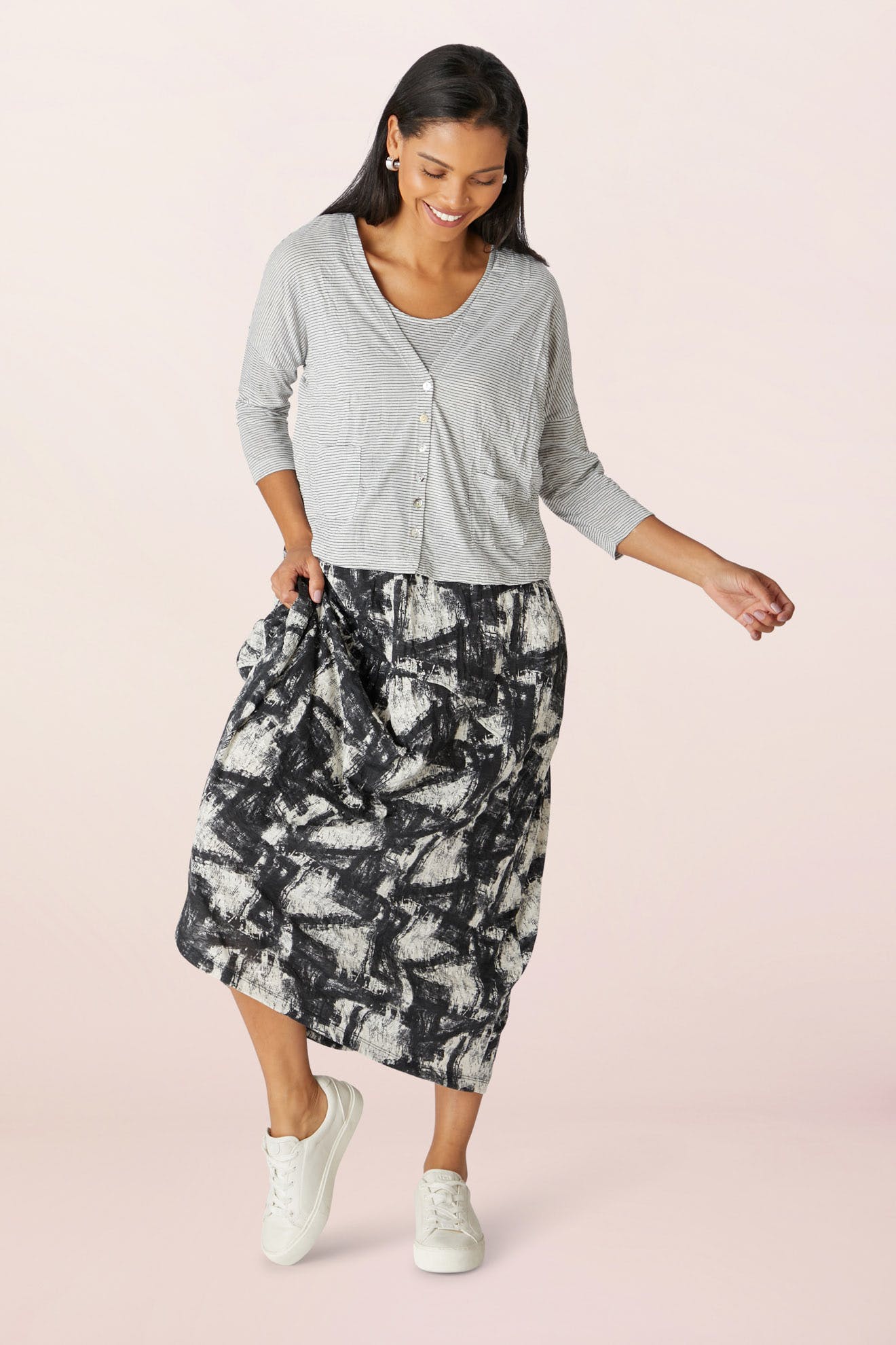 A woman in a Stripe Button Cardi and patterned skirt smiles as she walks, paired with white sneakers against a pink background.