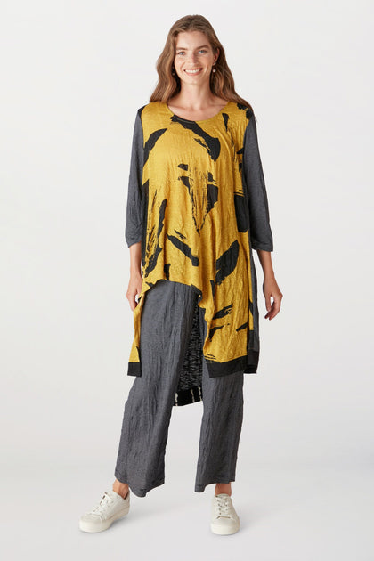 A woman smiling and posing in a trendy, layered outfit with a bold yellow and black tunic over gray Alembika Stripe Pants with an elasticated waist, complemented by white sneakers.