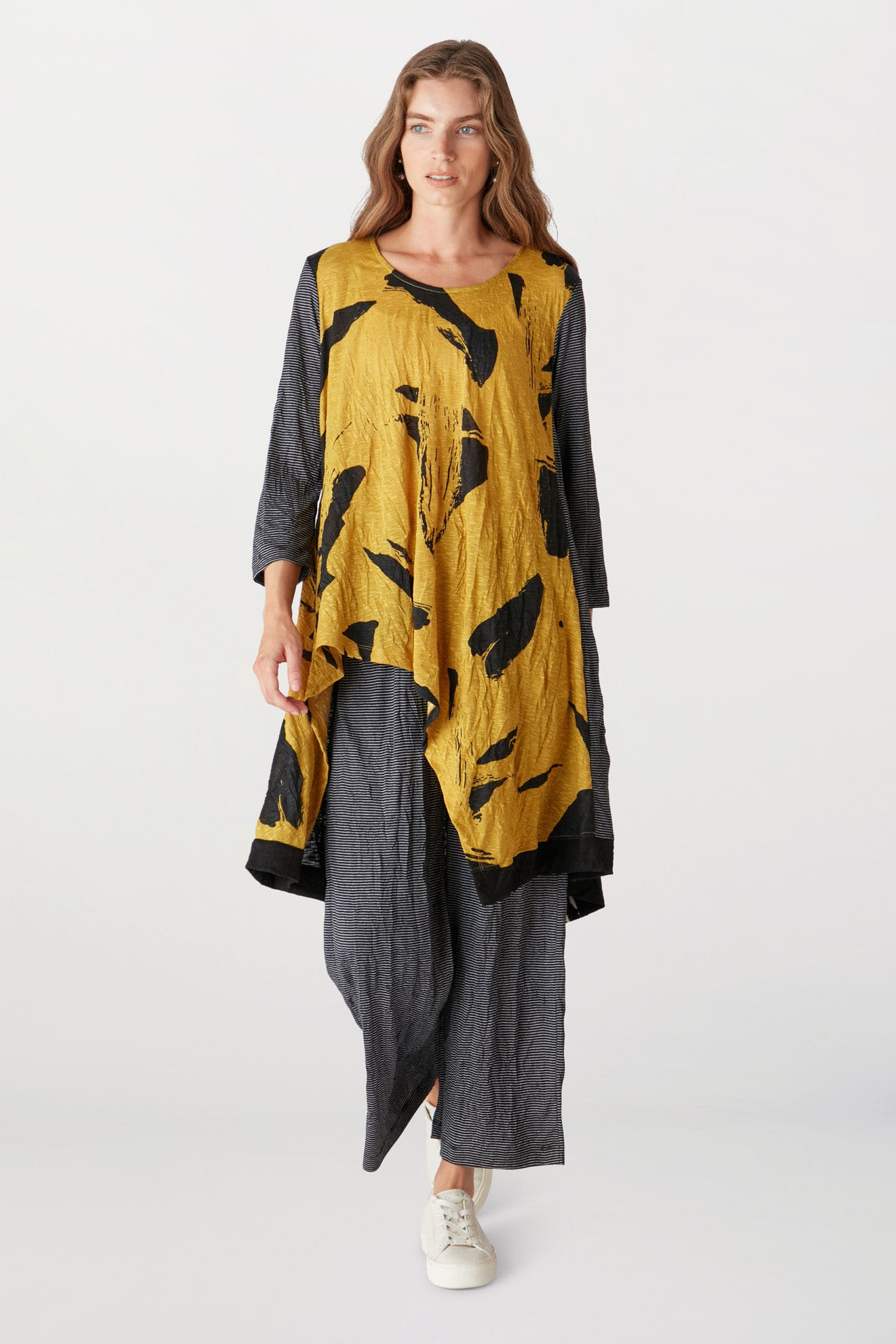 A woman stands wearing a stylish, asymmetric yellow and black dress with a gray Alembika underdress, paired with white sneakers.