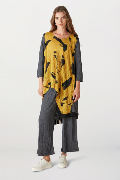 A woman in a studio wearing a yellow and black patterned tunic over grey Alembika Stripe Pants with an elasticated waist and white sneakers, standing against a plain background.