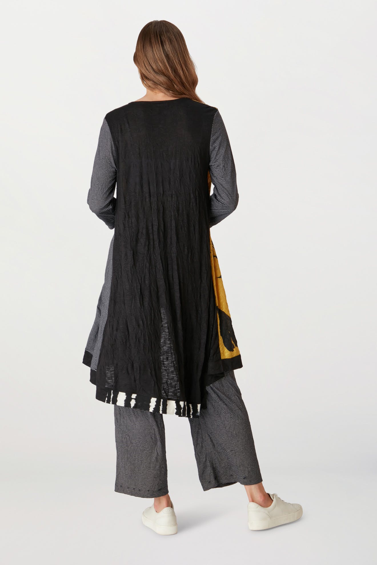 Woman in a trendy layered outfit, featuring a black crinkled jersey knit long jacket and gray Stripe Pant with an elasticated waist, viewed from the back on a plain background.