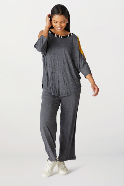 A woman stands smiling, wearing a casual gray outfit with a Cold Shoulder Top featuring yellow shoulder accents, made from crinkled jersey material, paired with white sneakers.