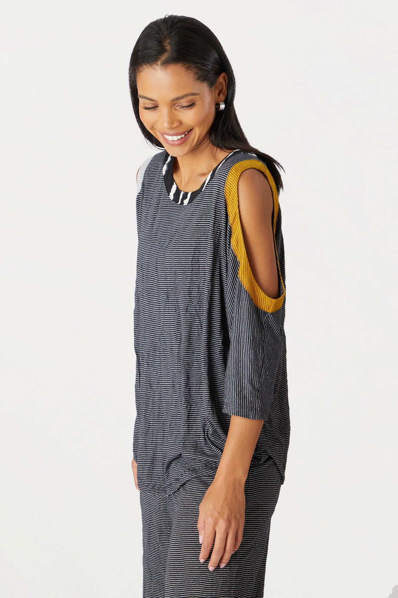 A woman smiling and looking down, wearing a striped yellow mesh side panel cold shoulder top, standing against a white background.