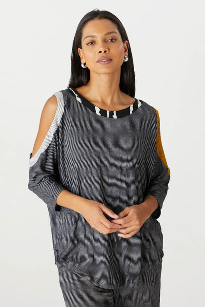 A woman stands facing the camera, wearing a stylish grey Alembika Cold Shoulder Top with cut-out shoulder details and a yellow stripe.