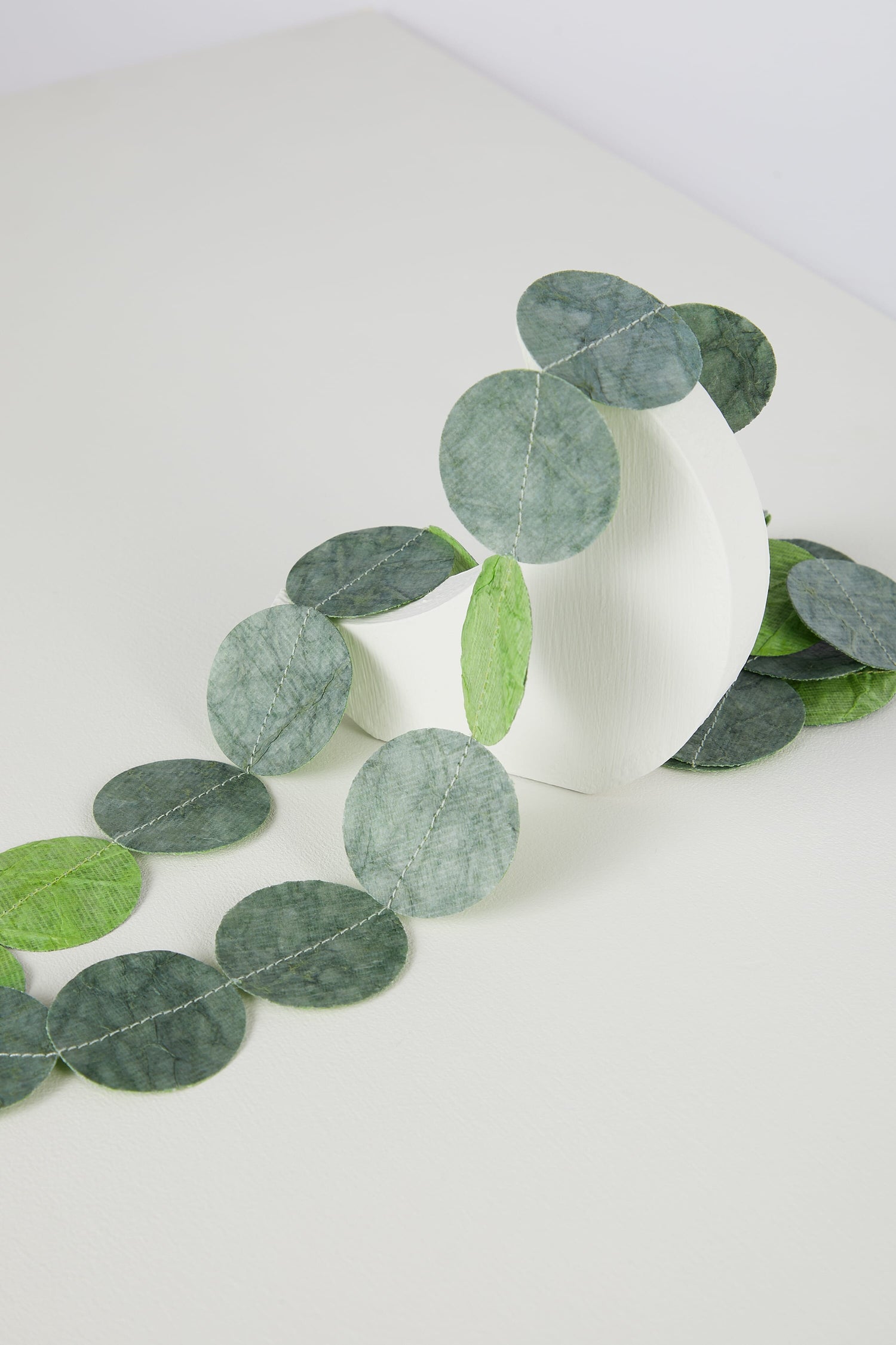 A white cylindrical Dotty Necklace with cascading delicate eucalyptus leaves on a white surface.