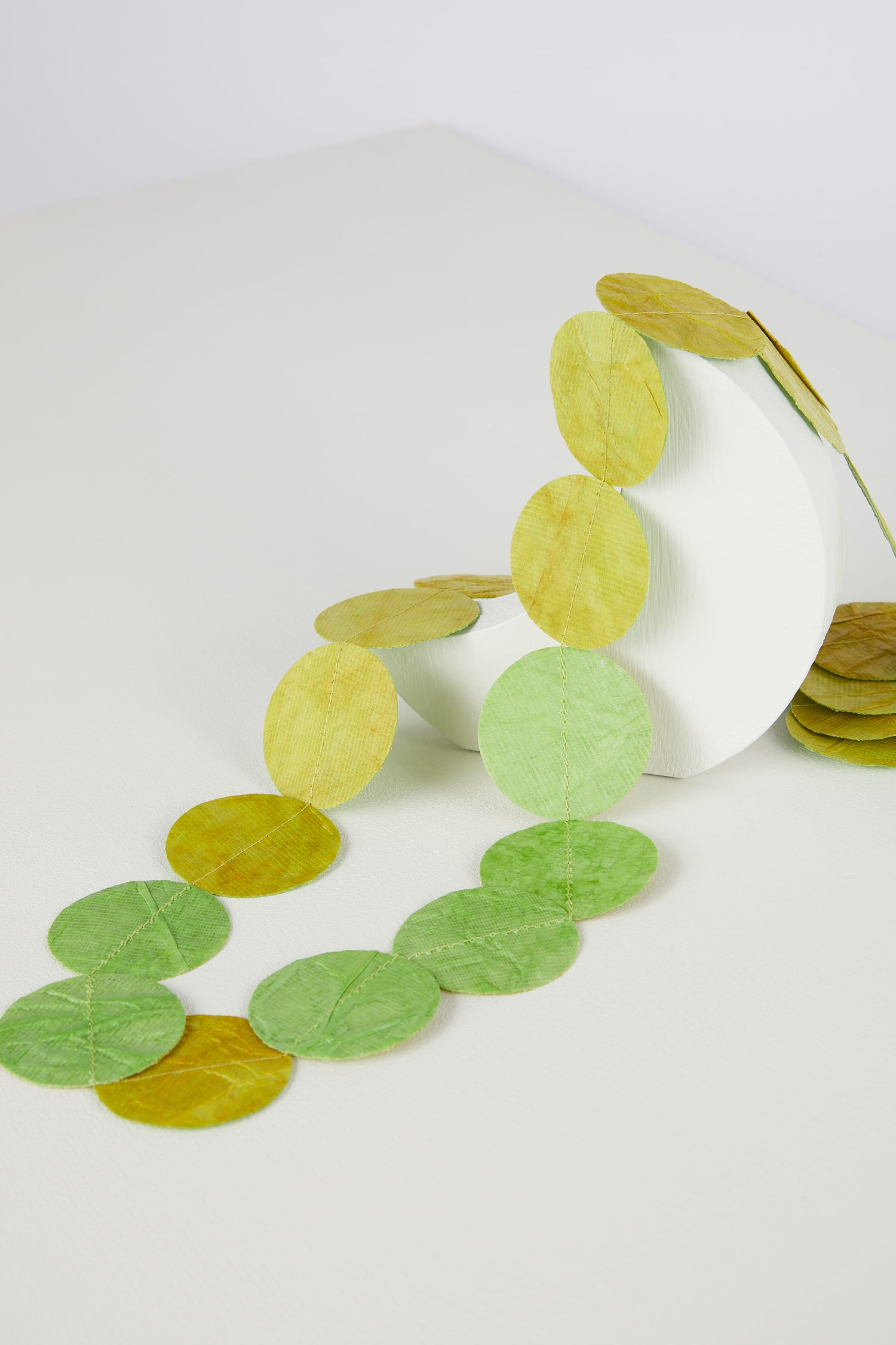 Transparent green and yellow hand-crafted leaf-shaped Dotty Necklace spilling out from a white cylindrical container on a desk.