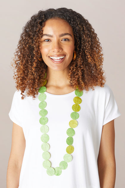 A smiling woman with curly hair wearing a white t-shirt, Dotty Necklace, and hoop earrings against a neutral background.