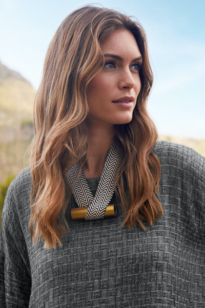Woman with long, wavy brown hair wearing a gray textured sweater and the Balance Necklace by Christina Brampti, looking to the side outdoors with mountains in the background.