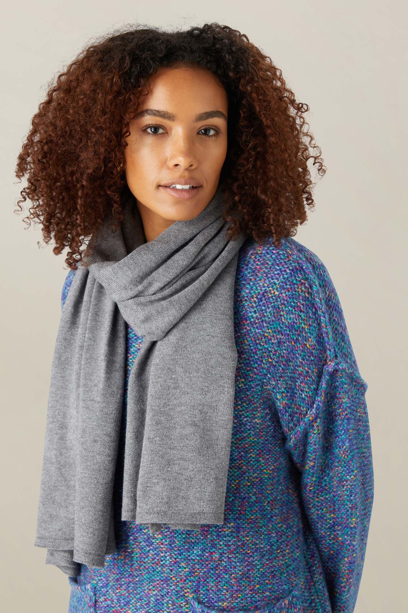 A person with curly hair wears the Knit Scarf in gray and a textured blue sweater, standing against a plain light background.
