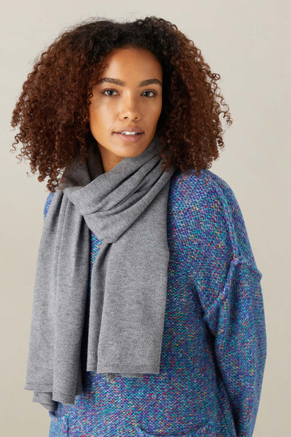 A person with curly hair wears the Knit Scarf in gray and a textured blue sweater, standing against a plain light background.