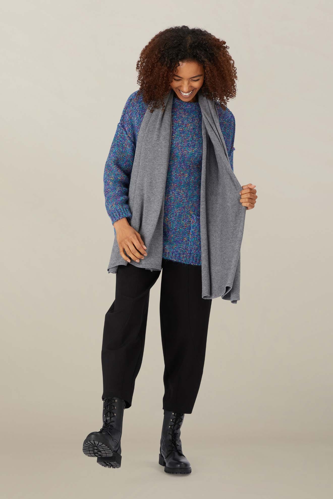 Person standing and smiling, wearing a blue sweater, Knit Scarf in gray, black pants, and black lace-up boots against a neutral background.