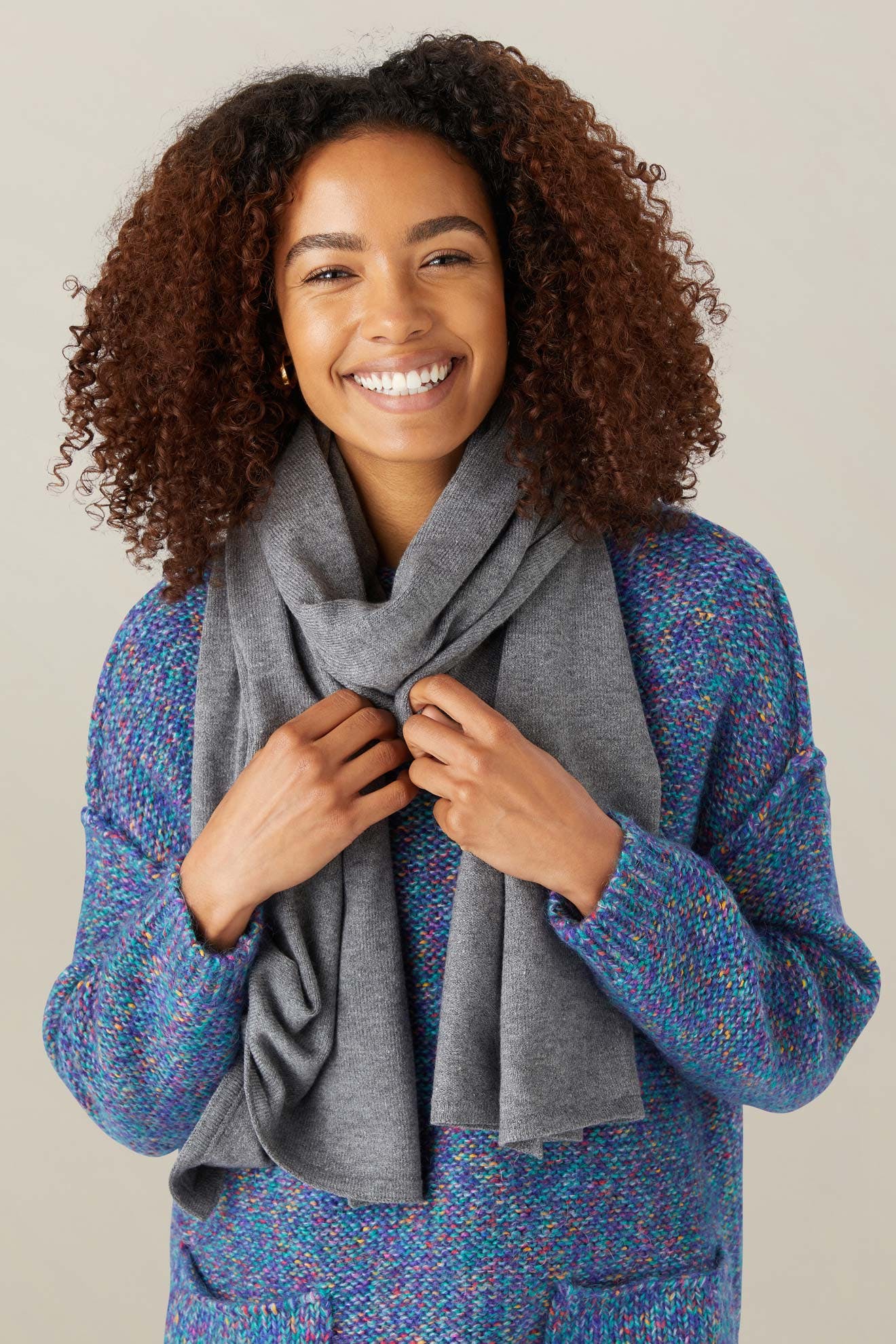 A woman with curly hair smiles at the camera, wearing a blue textured sweater and wrapping a Knit Scarf around her neck.