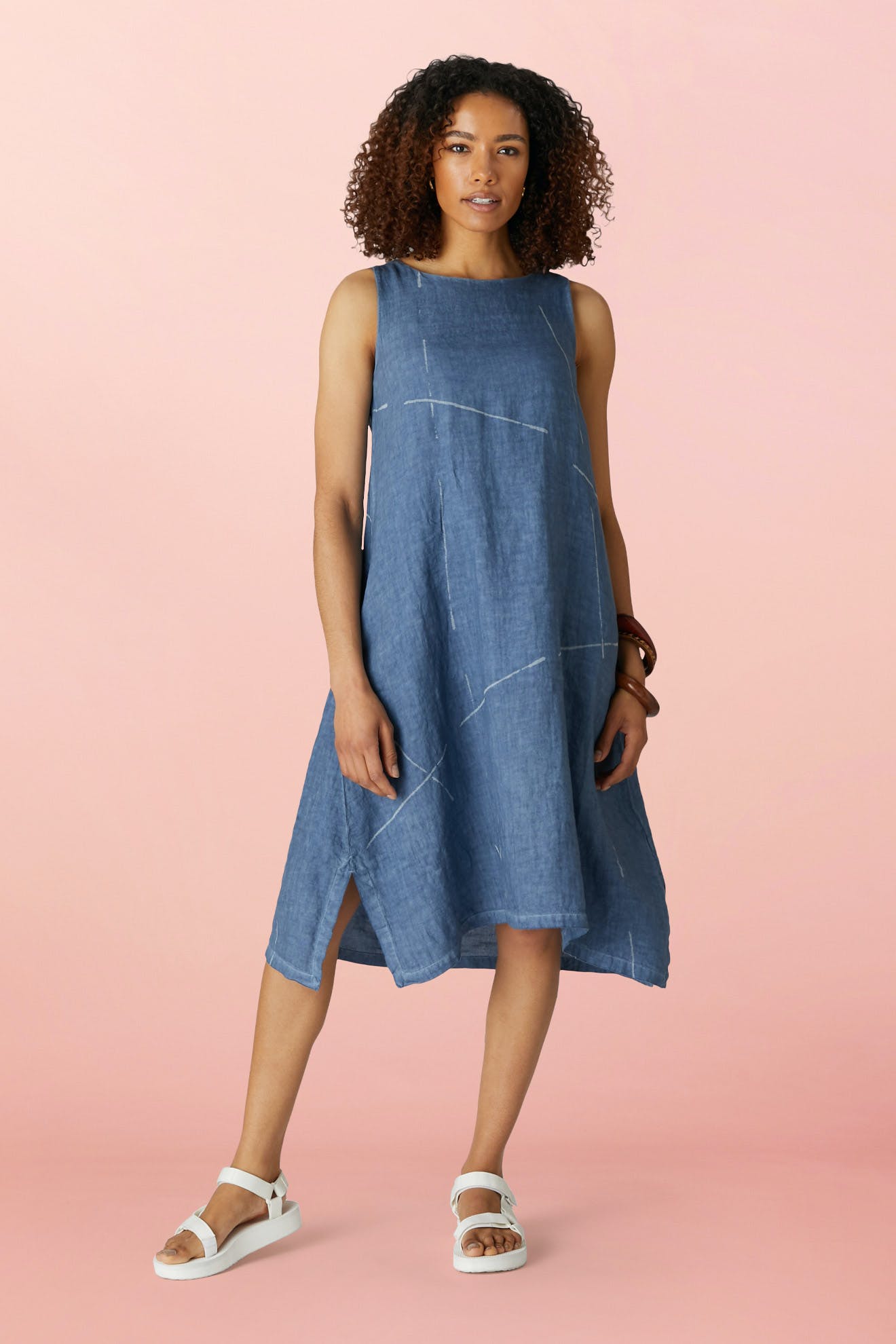 A woman stands against a pink background wearing the Som Linen Dress in blue with white lines, featuring a relaxed A-line silhouette, paired with white sandals.