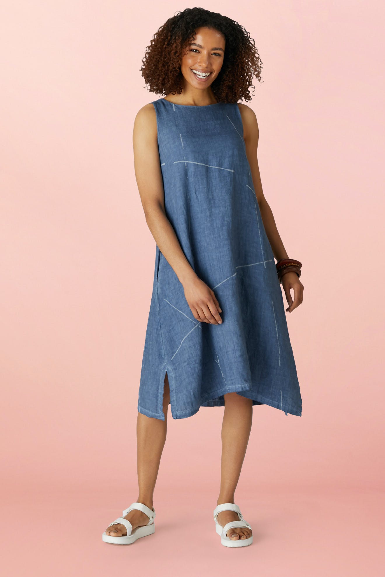 A woman smiles in the Som Linen Dress—a minimalist sleeveless design with a relaxed A-line silhouette and white stitching—paired with white sandals and a brown bracelet, set against a pink background.